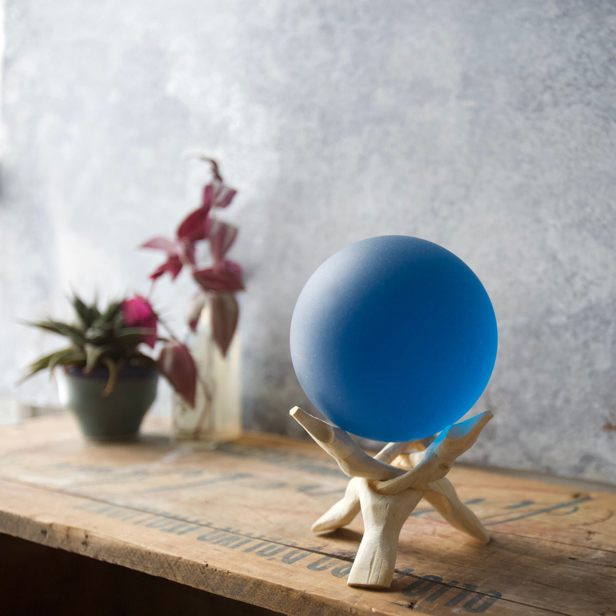 May include: A blue glass sphere on a wooden stand. The sphere is smooth and reflective. The stand is made of light brown wood and has three legs.