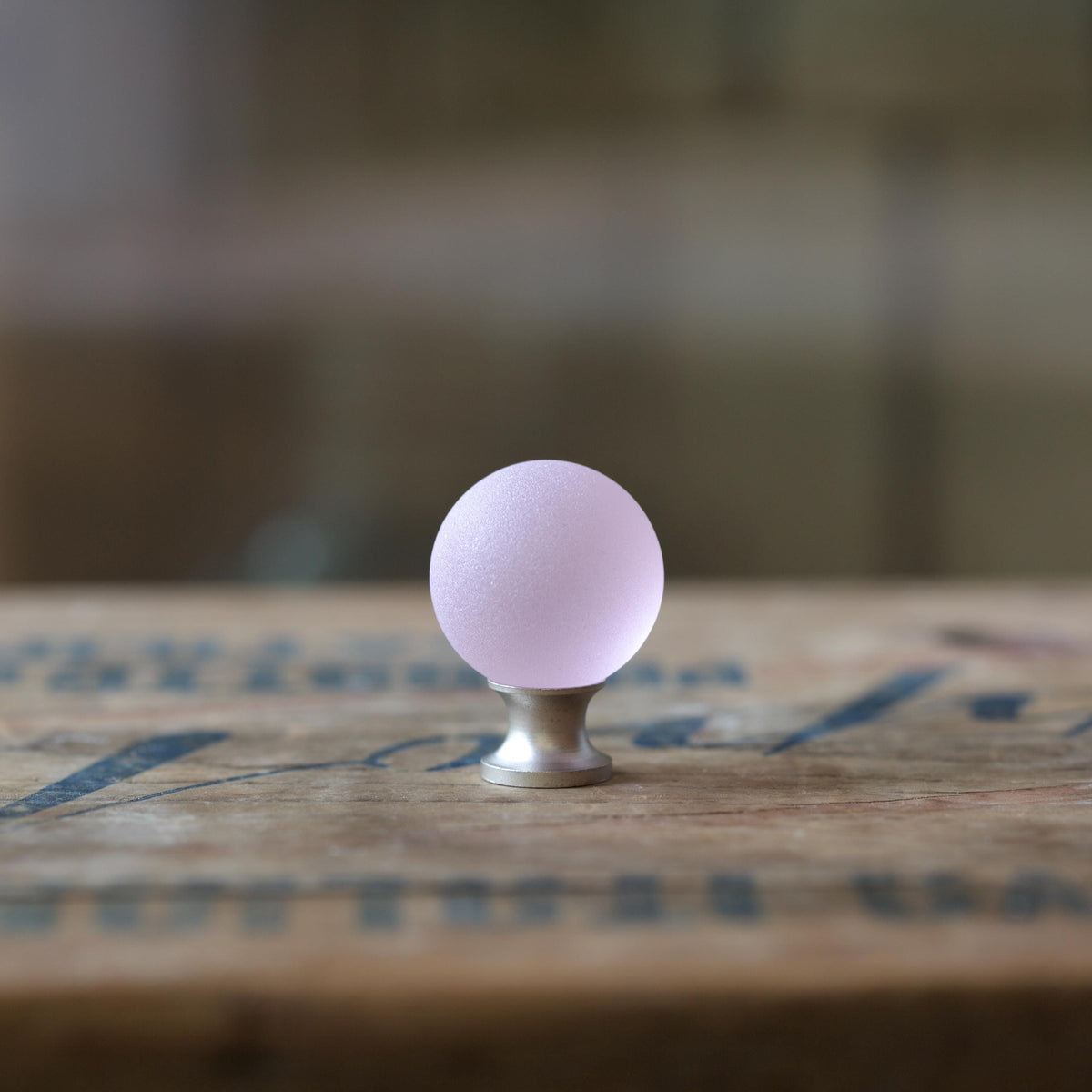 May include: Close-up view of a pale pink glass cabinet knob with a frosted finish. The knob is spherical and mounted on a small, silver-toned metal base. The background is blurred, suggesting a wooden surface.